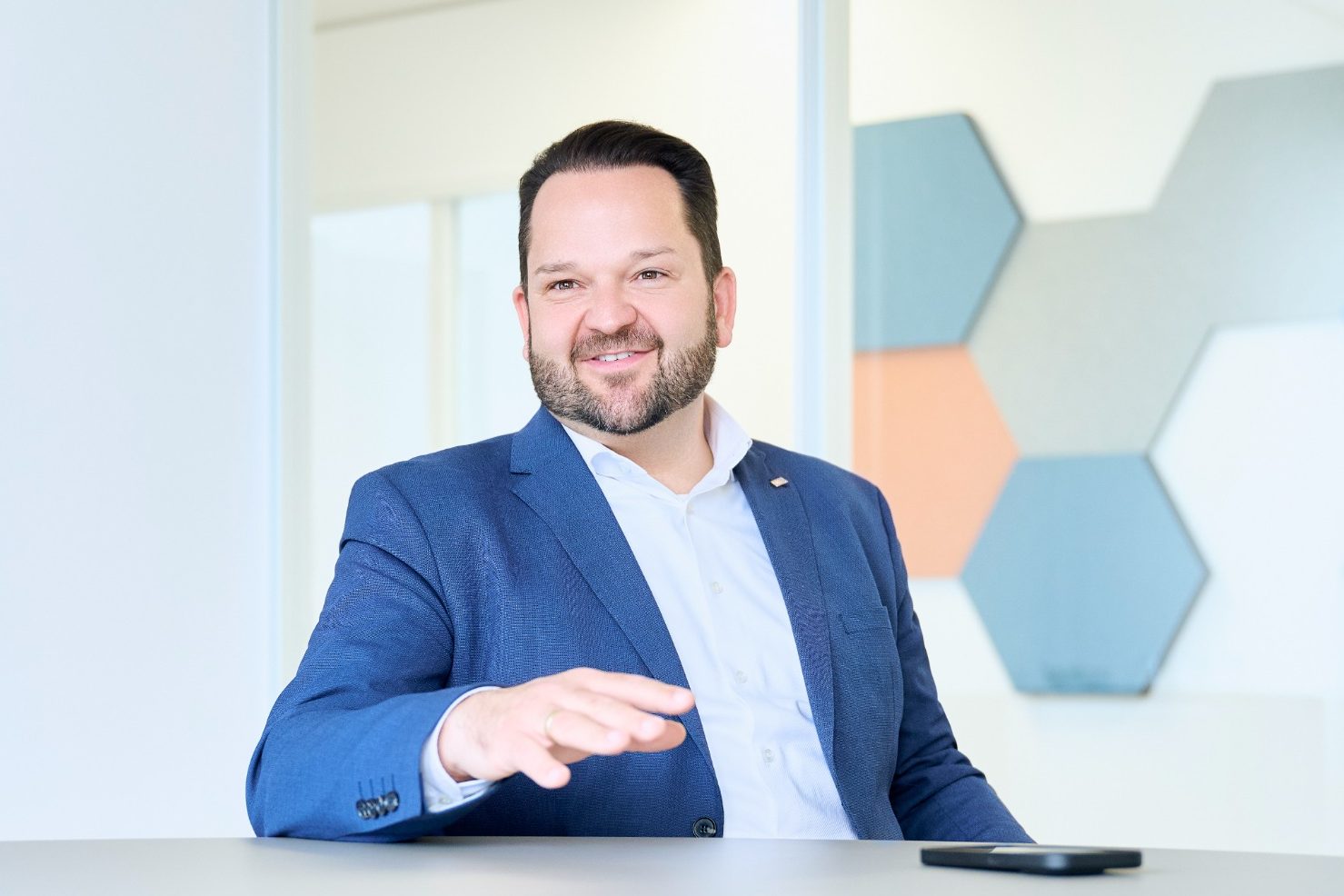 Mann mit Bart in formeller Kleidung, lächelt vor einem modernen Hintergrund.