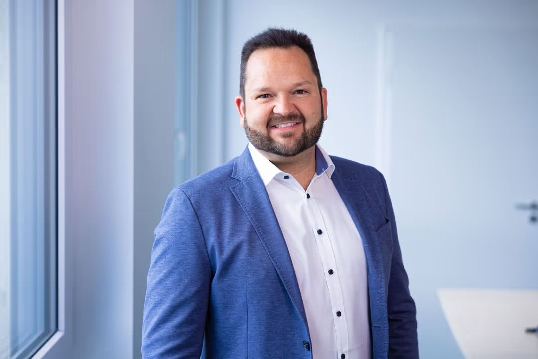 Timo Blei Mann mit Bart in formeller Kleidung, lächelt vor einem modernen Hintergrund.